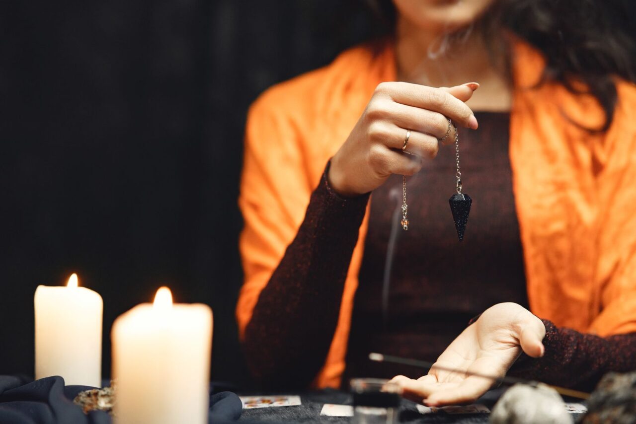 Mano sosteniendo un péndulo junto a velas encendidas